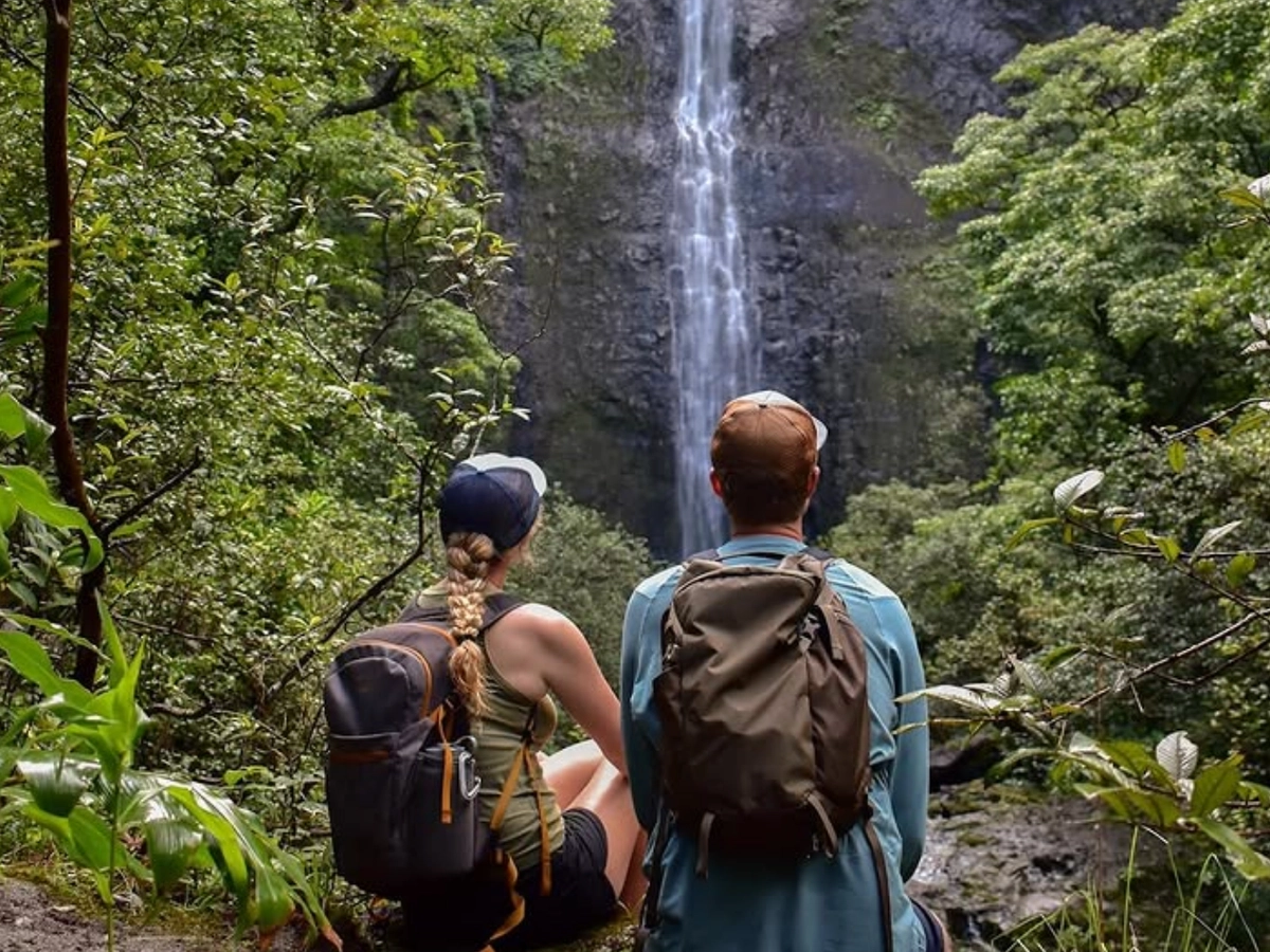 Two hikers with backpacks sit on a forest trail, gazing at a tall waterfall cascading down a rocky cliff surrounded by lush greenery.