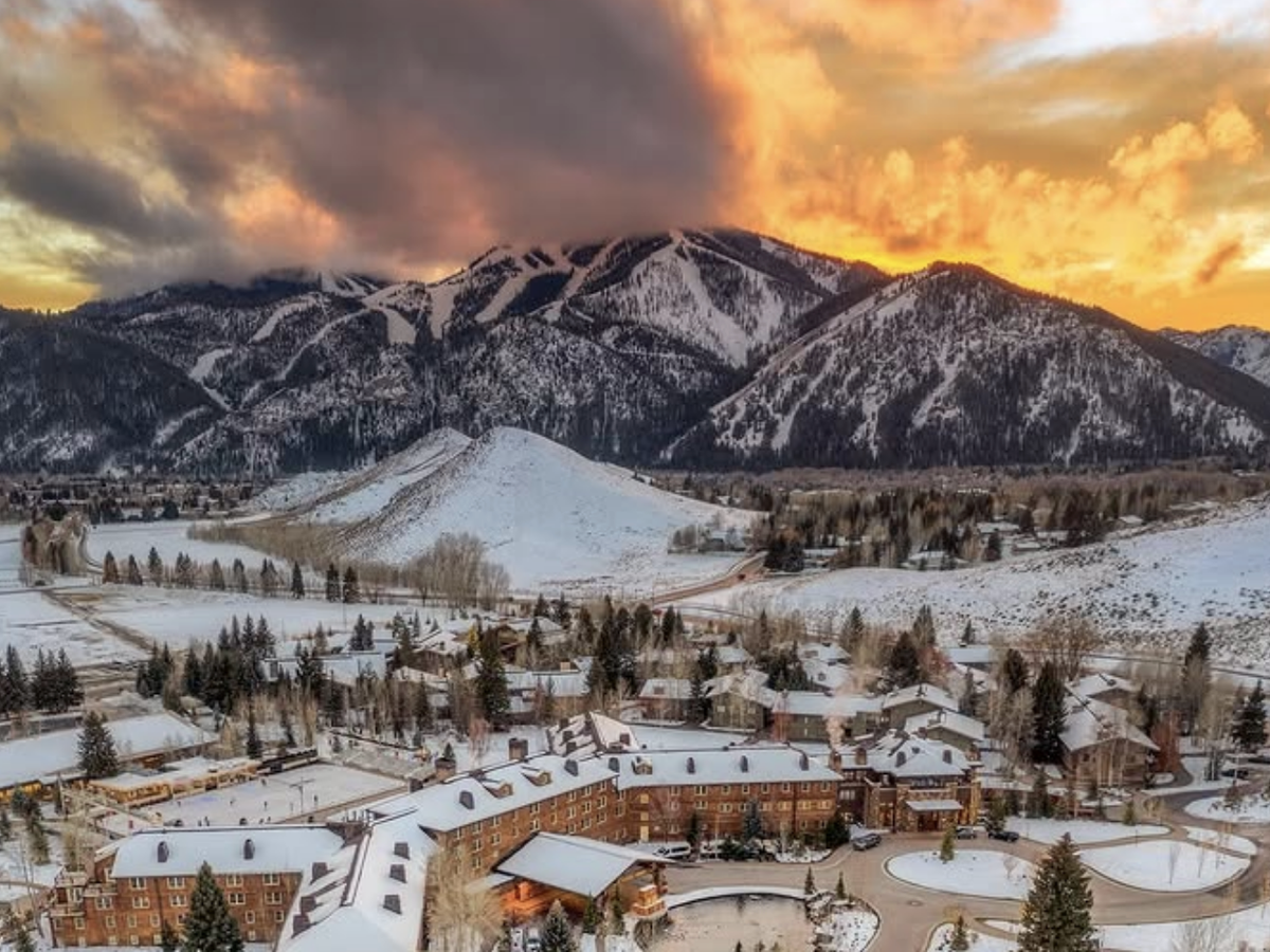 A winter resort town nestled at the base of snowy mountains glows under a dramatic orange and yellow sunset sky, with ski slopes visible in the distance.