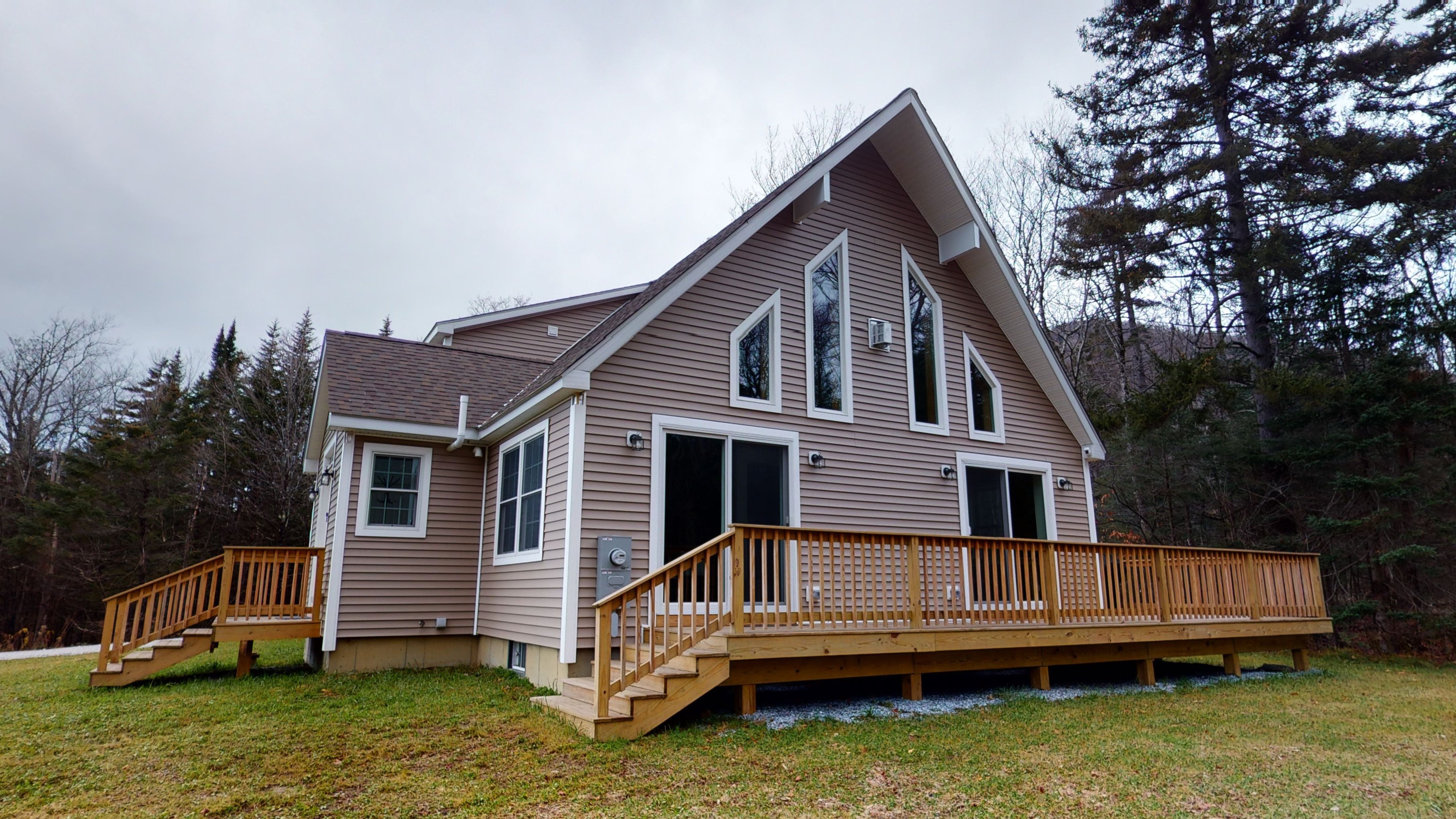 A modern A-frame-style cabin with beige siding and large triangular windows, featuring a spacious wooden deck and surrounded by a grassy yard and forest trees on an overcast day.