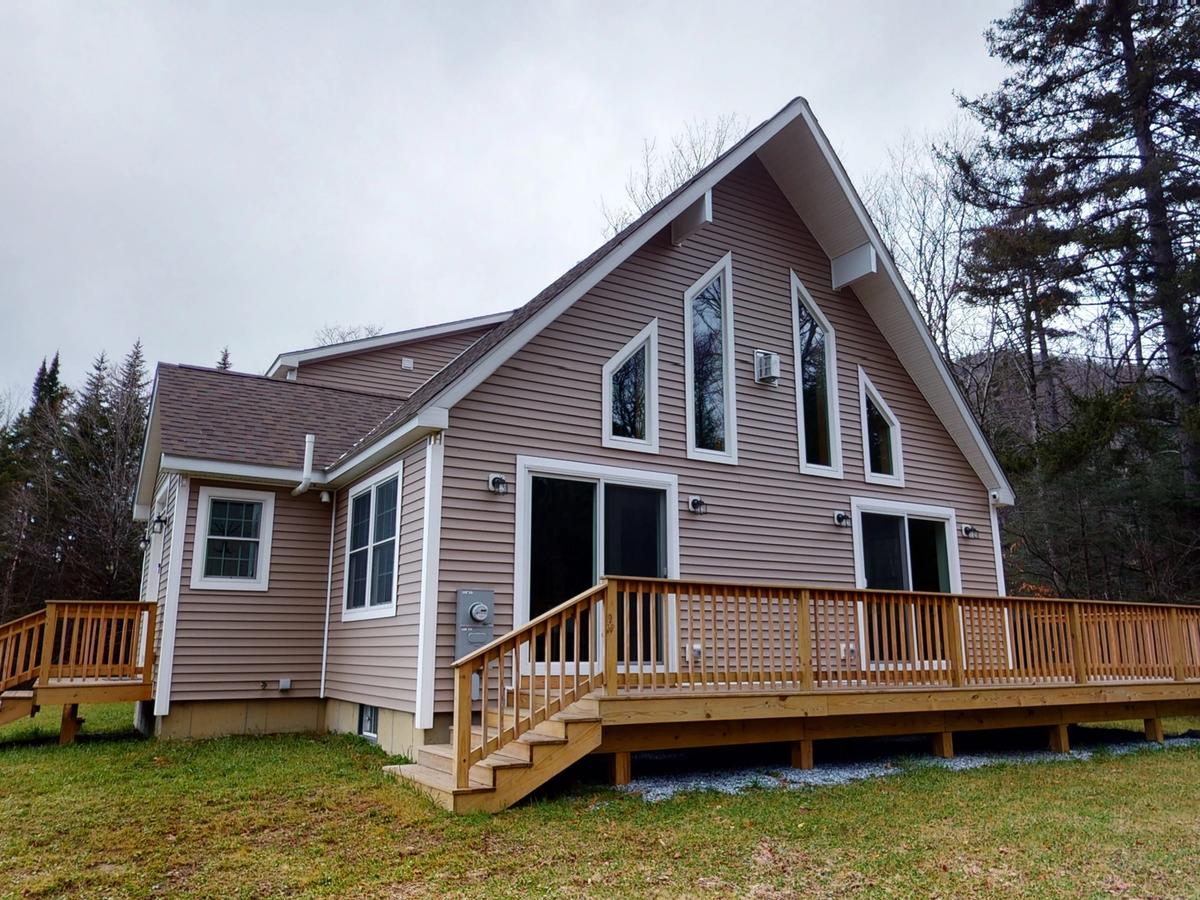 A modern A-frame-style cabin with beige siding and large triangular windows, featuring a spacious wooden deck and surrounded by a grassy yard and forest trees on an overcast day.