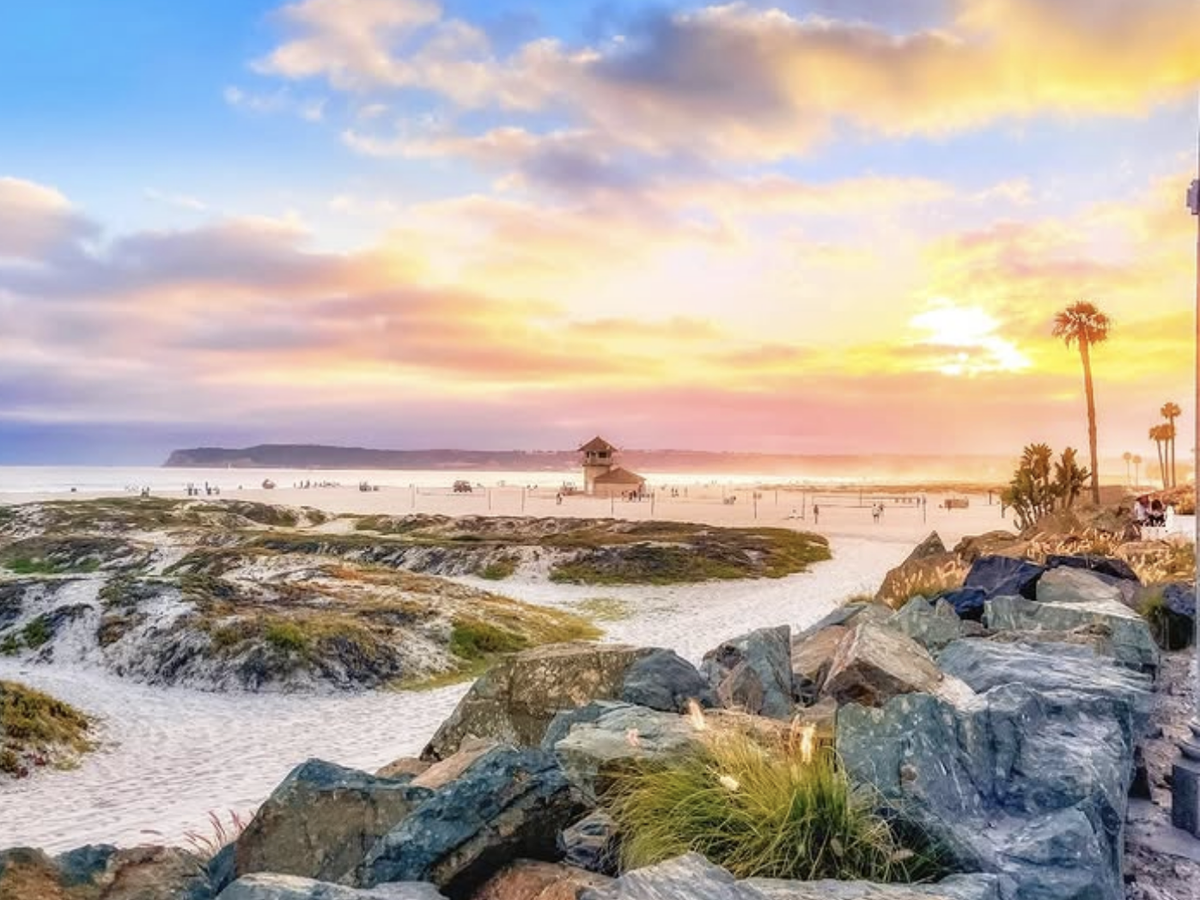 A colorful sunset over a sandy beach with rocky dunes, palm trees, and a lifeguard tower in the distance.