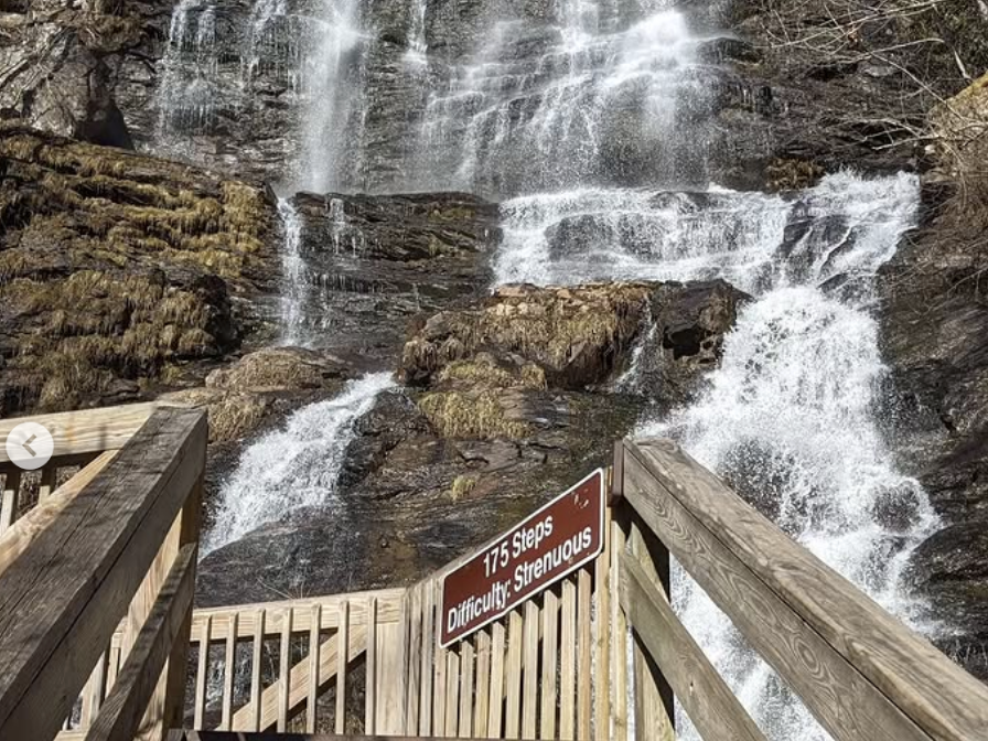 A steep wooden staircase leads up to a tall, cascading waterfall with a sign that reads “175 Steps, Difficulty: Strenuous” under a clear blue sky.