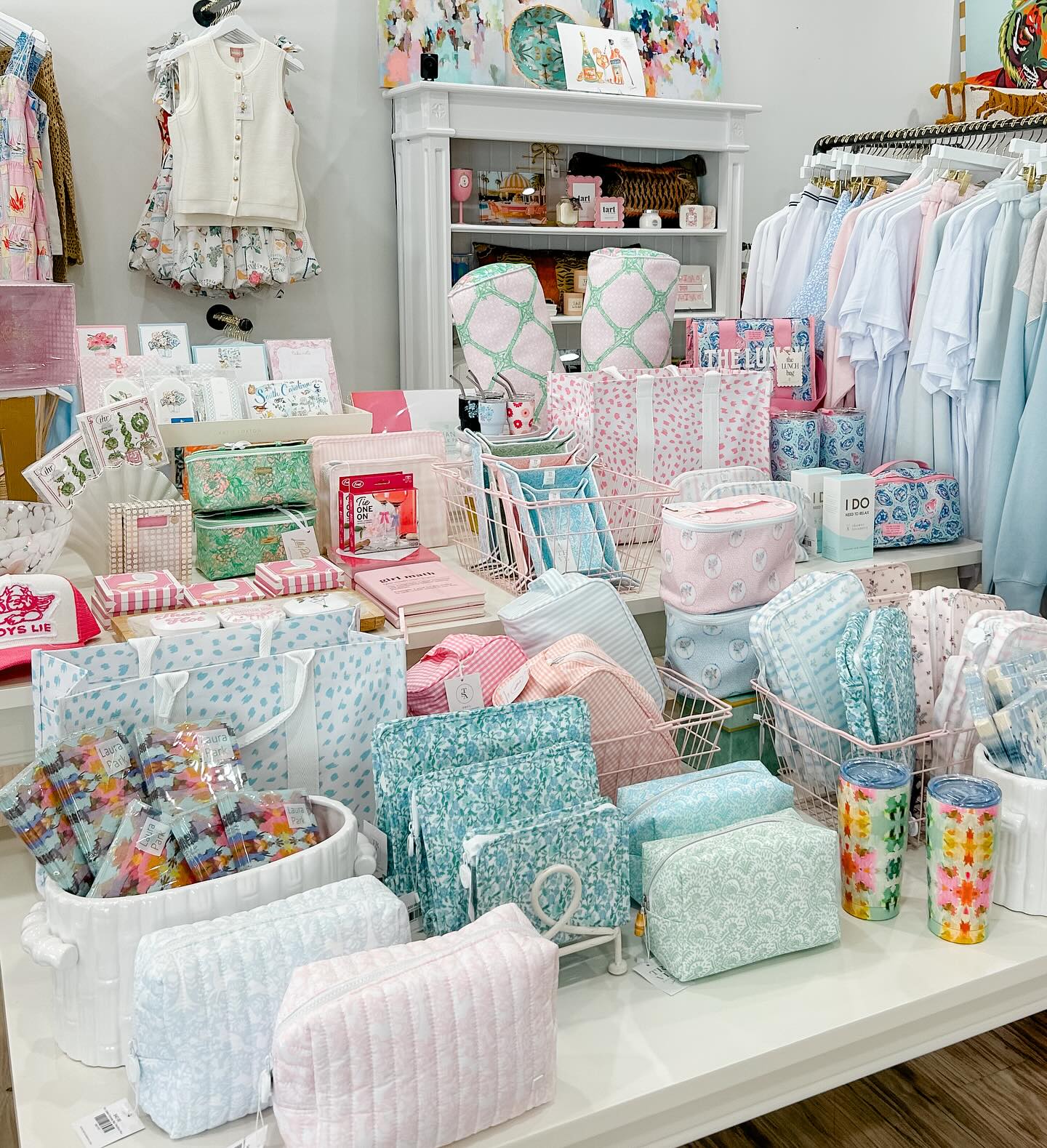 A display table at The Fizz Boutique overflows with pastel-colored cosmetic pouches, patterned tote bags, gift sets, stationery, and accessories in shades of mint, blush, and sky blue. Clothing racks with breezy tops and a floral children's outfit hang in the background alongside colorful wall art, creating a bright and feminine boutique atmosphere. 