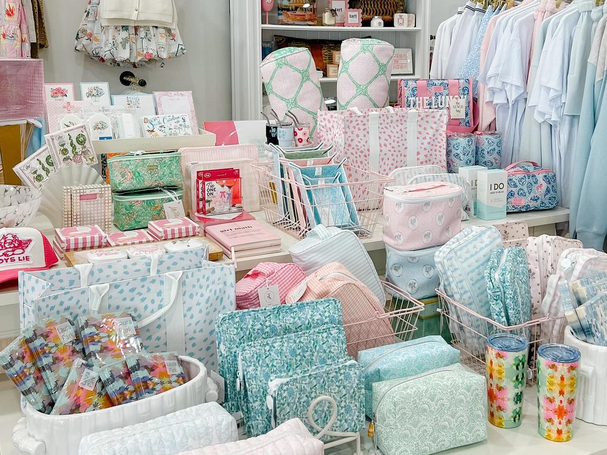 A display table at The Fizz Boutique overflows with pastel-colored cosmetic pouches, patterned tote bags, gift sets, stationery, and accessories in shades of mint, blush, and sky blue. Clothing racks with breezy tops and a floral children's outfit hang in the background alongside colorful wall art, creating a bright and feminine boutique atmosphere.