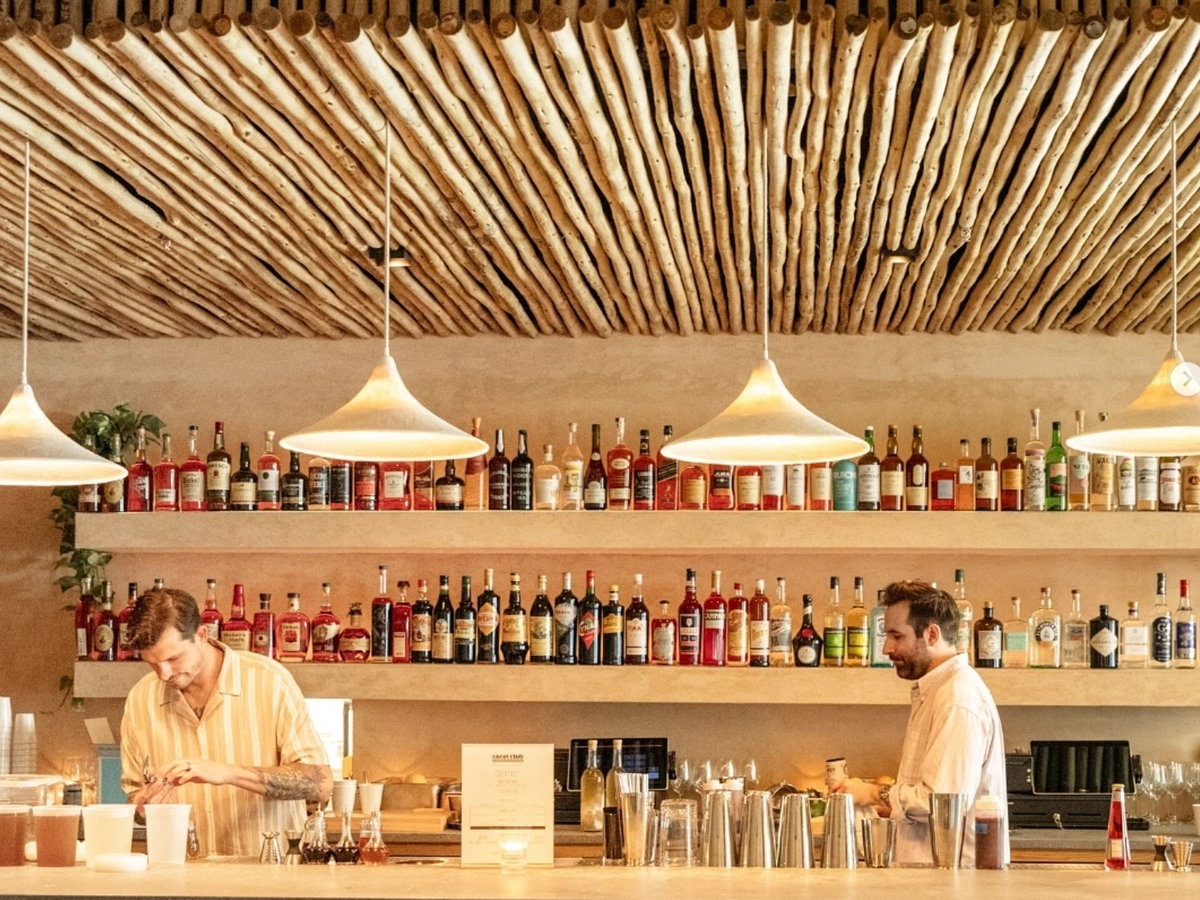A stylish bar with warm lighting, natural wood accents, and shelves lined with colorful liquor bottles as two bartenders prepare drinks.