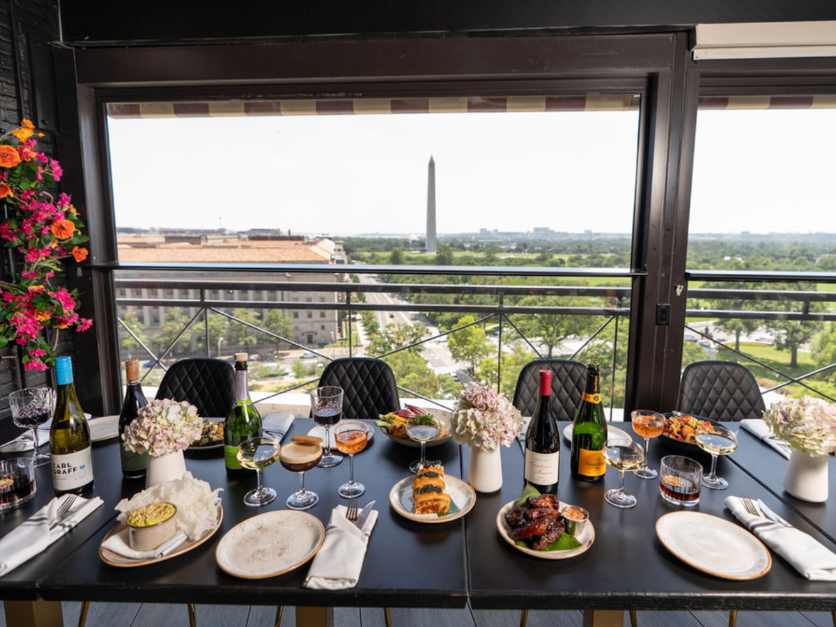 Elegant rooftop dining table set with wine, cocktails, and plated dishes, overlooking the Washington Monument and cityscape in the distance.