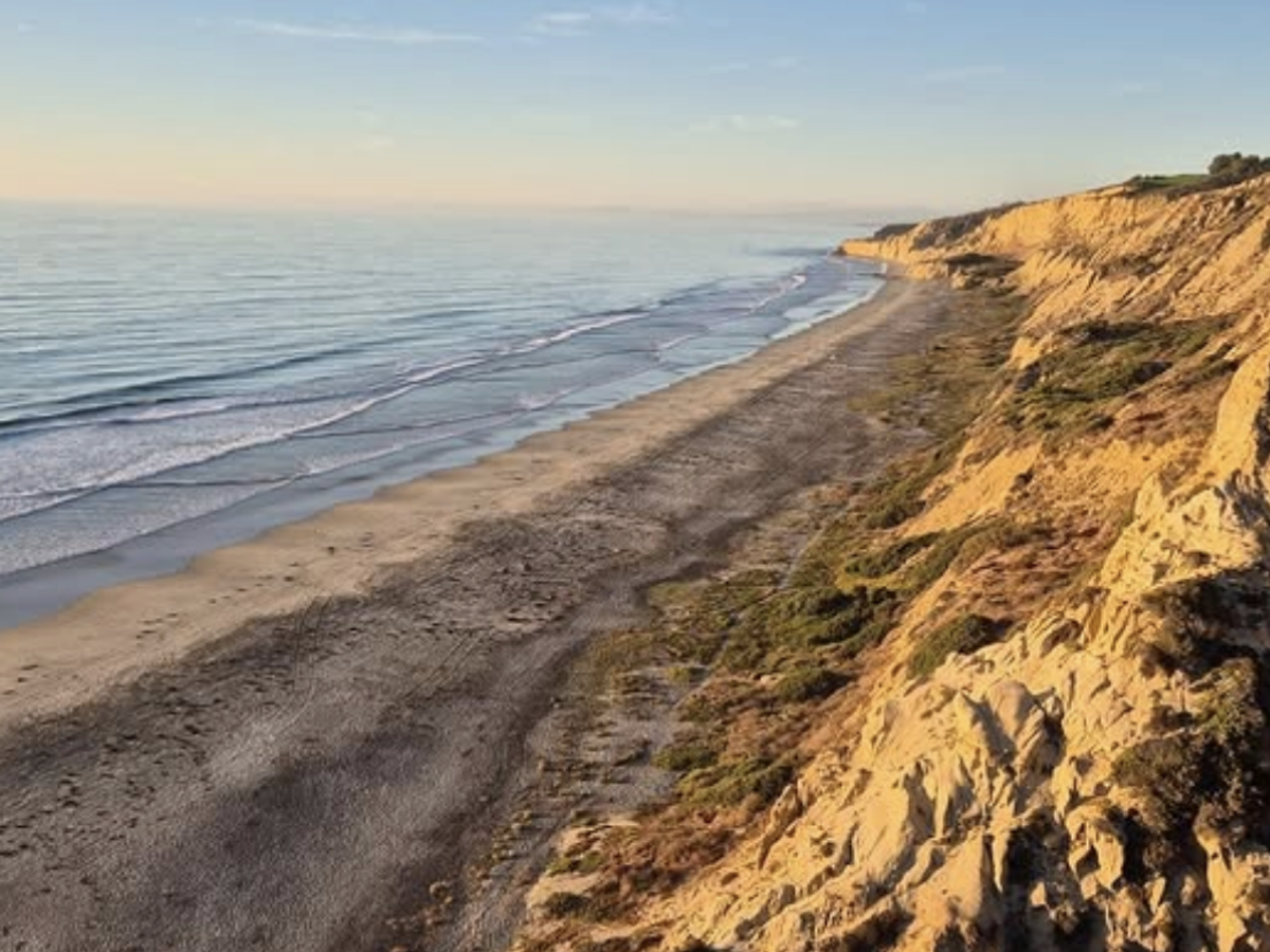Two people walk along a quiet beach tucked between tall cliffs as soft sunlight shines through the coastal haze. This hidden stretch of sand shows the calm and peaceful side of secluded beaches in San Diego.