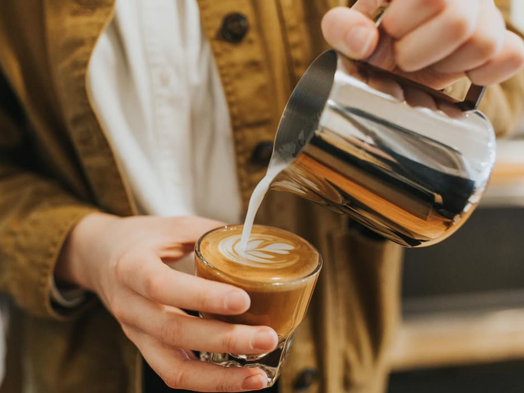 A barista wearing a brown jacket pours steamed milk from a stainless steel pitcher into a small glass, creating intricate latte art on a cortado. The close-up shot captures the precision of the pour and the rich texture of the coffee in a warmly lit café setting.