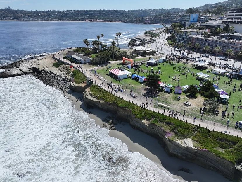 An aerial view of La Jolla’s coastline, showing a green park filled with booths, ocean cliffs, and crashing waves. It’s the perfect mix of seaside fun and community events.