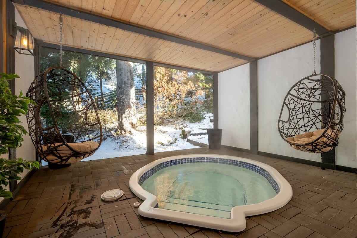 An indoor soaking pool with hanging wicker chairs creates a cozy spa-like space at Chalet Whistler, with large windows showing snowy trees outside. This relaxing indoor retreat is ideal for guests who want a warm place to unwind after a day of mountain adventures.