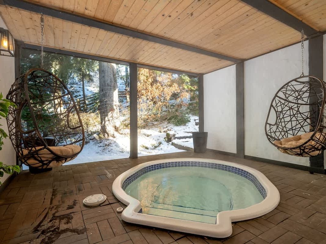 An indoor soaking pool with hanging wicker chairs creates a cozy spa-like space at Chalet Whistler, with large windows showing snowy trees outside. This relaxing indoor retreat is ideal for guests who want a warm place to unwind after a day of mountain adventures.
