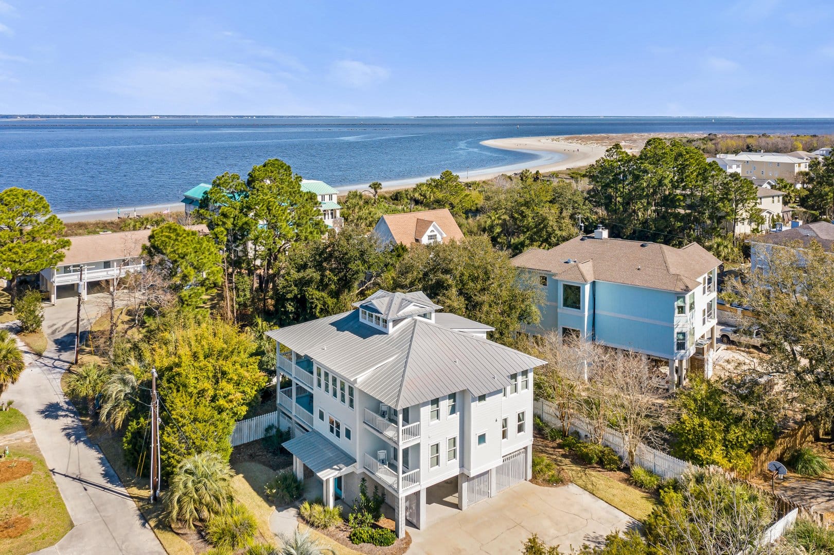 A coastal vacation home with a modern, light gray exterior and a metal roof, surrounded by lush greenery and neighboring beach houses. The elevated design includes multiple balconies and a covered carport. The background features a serene shoreline with a curved sandbar and calm blue waters stretching to the horizon