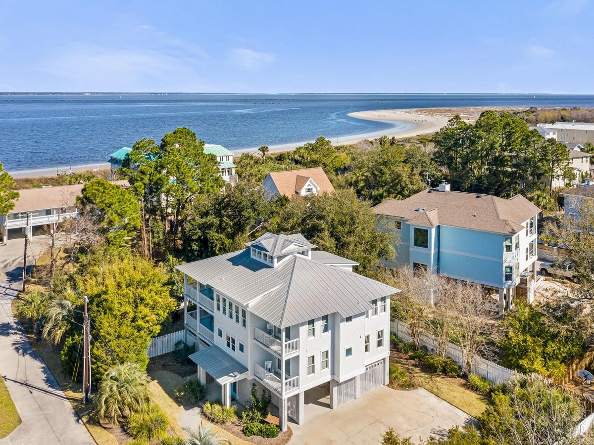 A coastal vacation home with a modern, light gray exterior and a metal roof, surrounded by lush greenery and neighboring beach houses. The elevated design includes multiple balconies and a covered carport. The background features a serene shoreline with a curved sandbar and calm blue waters stretching to the horizon