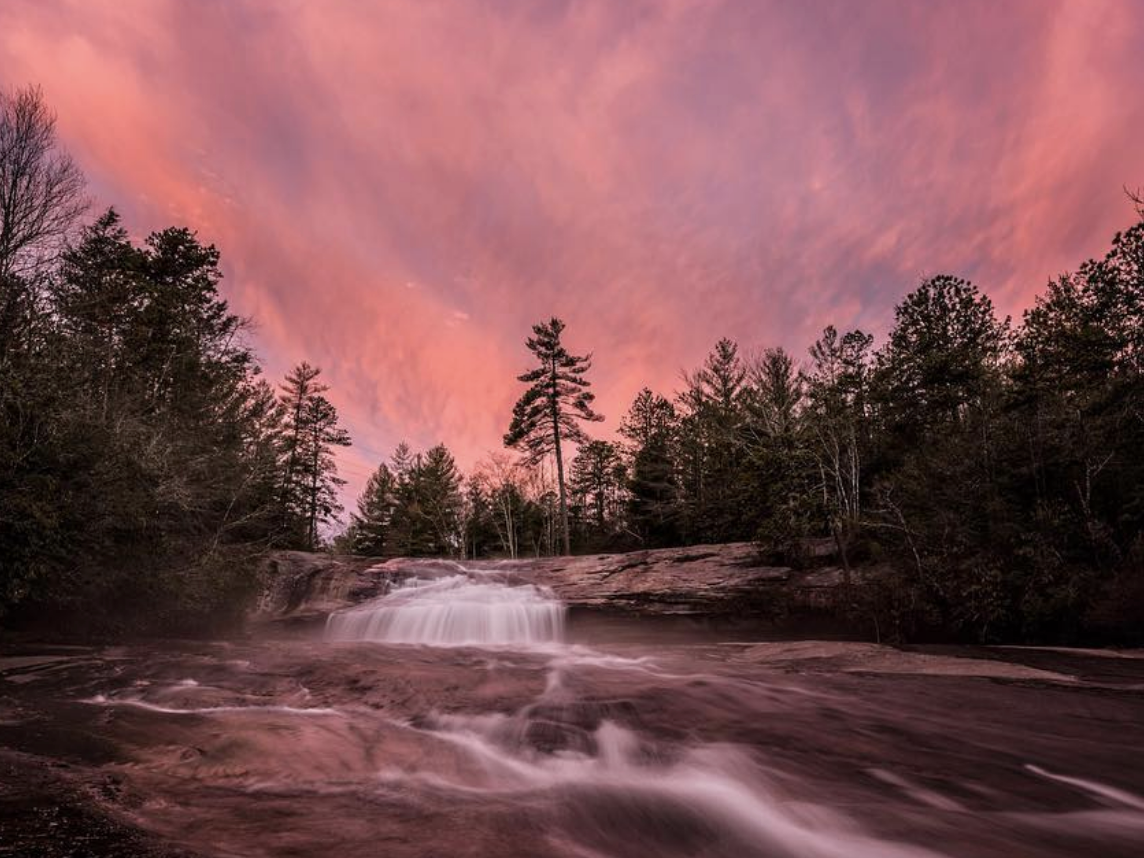 A cascading waterfall flows over smooth rock formations, captured with a silky effect, beneath a dramatic pink and purple sunset sky. The surrounding forest of tall pine trees and dense foliage frames the scene, creating a serene and mystical atmosphere.