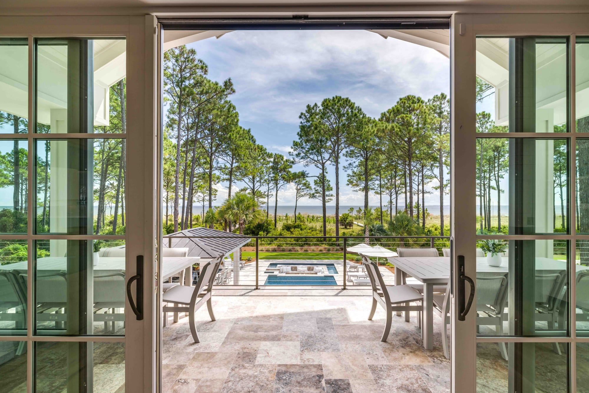 Through large glass doors, you see a peaceful patio with tall pine trees and water beyond. Outdoor seating and a pool create a private, resort-like vibe. It’s a calm place to enjoy quiet mornings or sunset views.