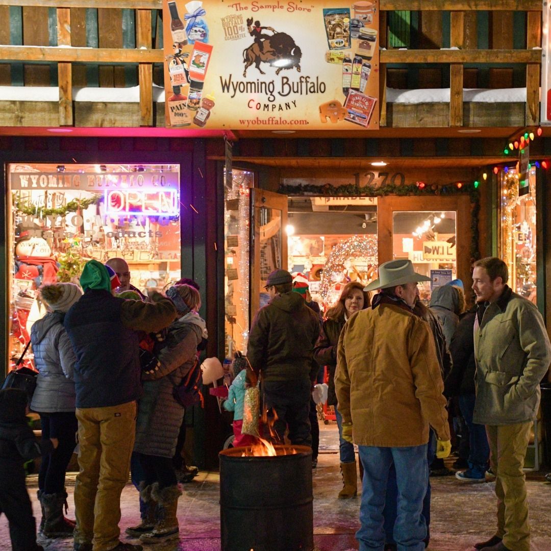 Locals gather outside the Wyoming Buffalo Company store, warming up by a fire barrel as holiday lights twinkle around them. The festive crowd laughs and mingles, capturing the cozy spirit of a small-town Christmas night.