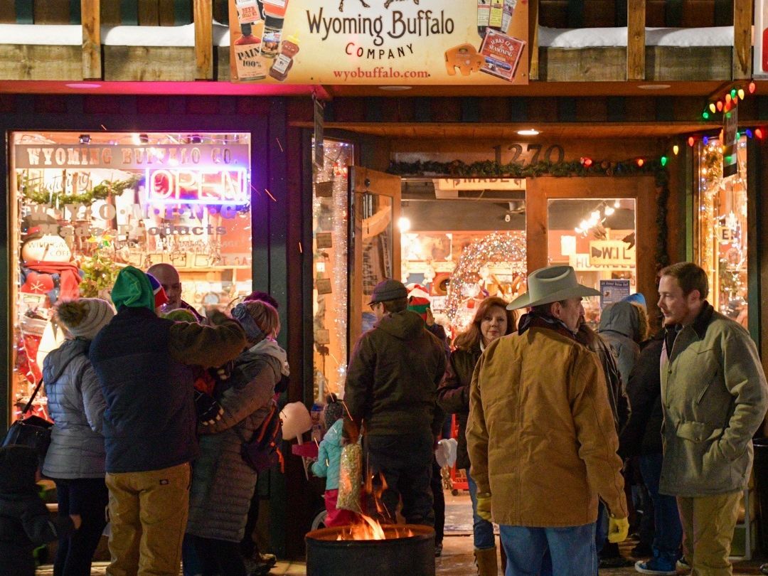 Locals gather outside the Wyoming Buffalo Company store, warming up by a fire barrel as holiday lights twinkle around them. The festive crowd laughs and mingles, capturing the cozy spirit of a small-town Christmas night.