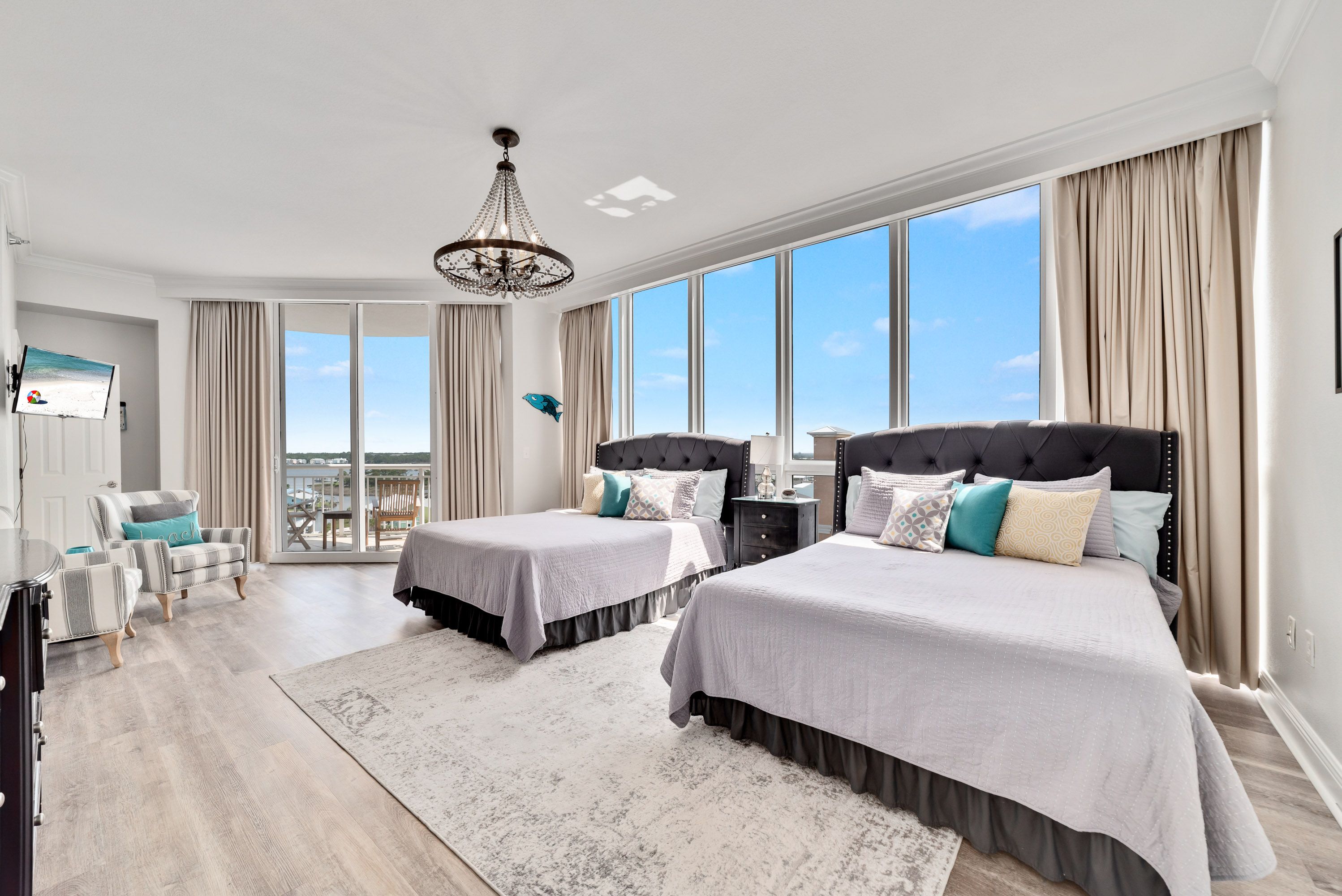 A bright and spacious coastal bedroom with two double beds, tufted headboards, elegant pillows, a chandelier, floor-to-ceiling windows, and a private balcony overlooking water views.