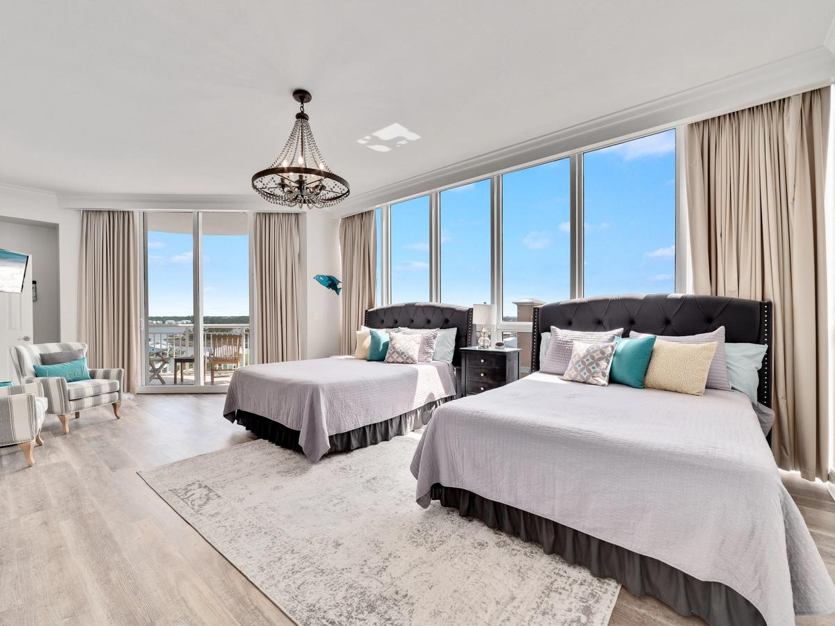 A bright and spacious coastal bedroom with two double beds, tufted headboards, elegant pillows, a chandelier, floor-to-ceiling windows, and a private balcony overlooking water views.