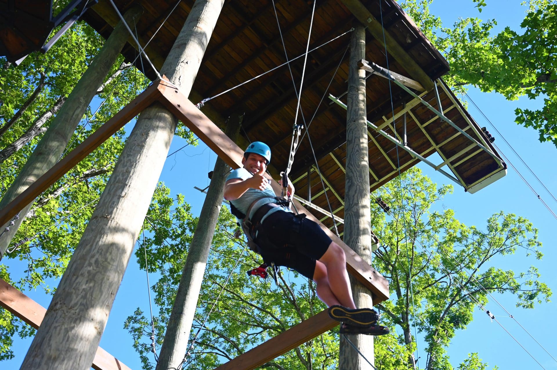 A smiling adventurer dangles mid-air on a dueling zipline course at Anakeesta surrounded by tall trees. The ropes and platforms blend into the lush forest, making it an exciting treetop challenge.