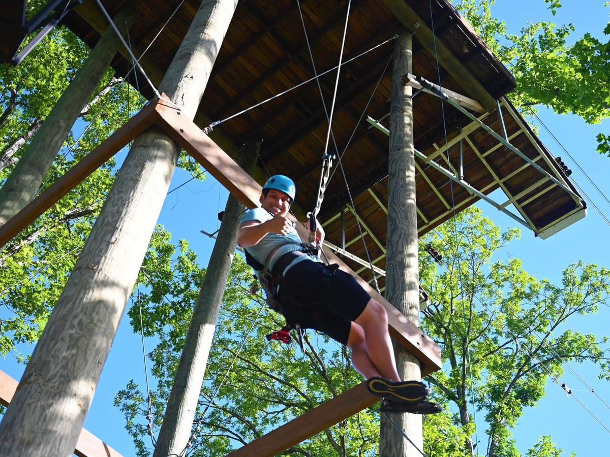 A smiling adventurer dangles mid-air on a dueling zipline course at Anakeesta surrounded by tall trees. The ropes and platforms blend into the lush forest, making it an exciting treetop challenge.