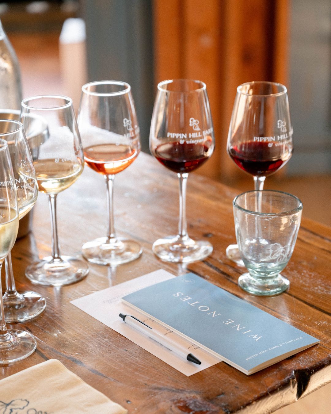 A wine tasting setup with several glasses of red, rosé, and white wine lined up on a rustic wooden table. A tasting booklet and pen sit nearby, showing the hands-on experience of learning about different wine styles. This scene reflects a casual yet thoughtful winery tasting that’s perfect for weekend visitors.