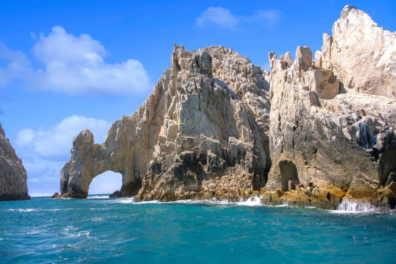 El Arco rock formation rises from the turquoise waters in Los Cabos, Mexico, with dramatic cliffs and a natural stone arch along the Pacific coast. Waves crash against the rugged shoreline under a bright blue sky, creating a stunning coastal view. This iconic landmark makes Los Cabos a top destination for beach lovers and ocean tours.