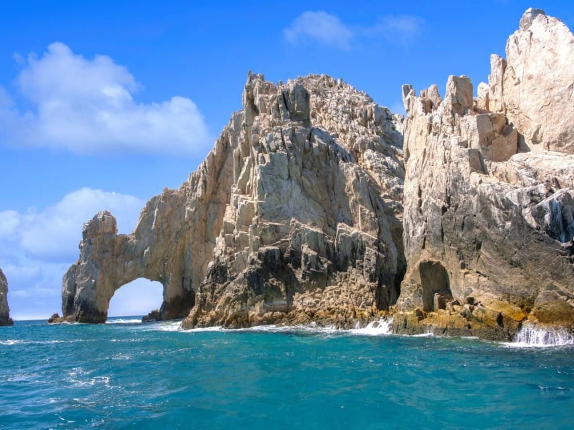 El Arco rock formation rises from the turquoise waters in Los Cabos, Mexico, with dramatic cliffs and a natural stone arch along the Pacific coast. Waves crash against the rugged shoreline under a bright blue sky, creating a stunning coastal view. This iconic landmark makes Los Cabos a top destination for beach lovers and ocean tours.