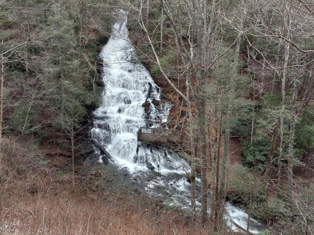 A scenic waterfall flows through Vogel State Park, surrounded by tall trees and a quiet forest setting. A wooden viewing platform sits beside the rushing water, giving visitors a peaceful spot to stop and enjoy nature. This area is popular for hiking, sightseeing, and relaxing outdoor escapes in North Georgia.