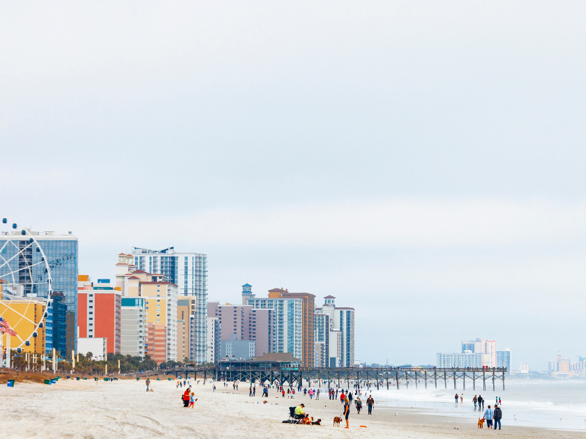 People stroll along the sandy shore of Myrtle Beach with the SkyWheel and high-rise hotels in the background. A calm day with soft clouds sets the scene for a classic beach town vibe along the South Carolina coast.