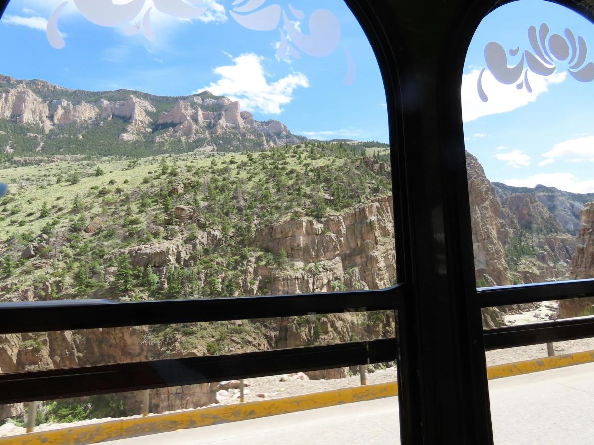 Rugged cliffs and pine-covered slopes fill the view from a vehicle window, with a steep canyon and sunlit mountains unfolding under a vivid blue Wyoming sky.