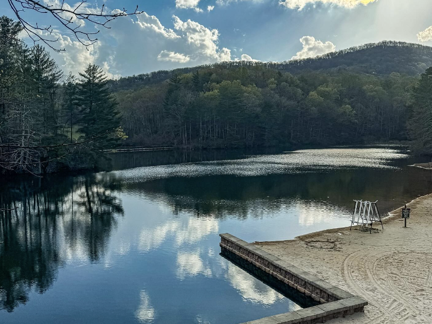A quiet lake bordered by sandy shoreline and forested hills, with reflections of clouds and trees on the calm water under a partly cloudy sky.