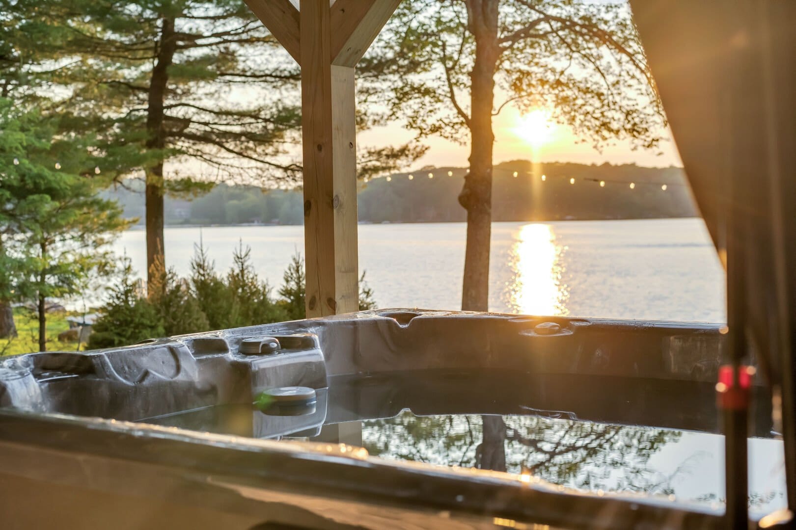 Covered outdoor hot tub overlooking a tranquil lake at sunset, with golden light reflecting on the water through surrounding trees, creating a peaceful, romantic cabin retreat atmosphere.