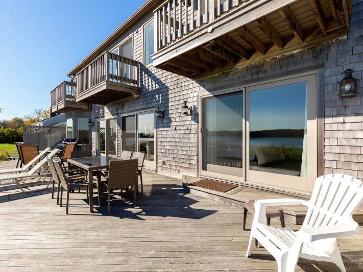 Spacious waterfront deck with weathered wood siding, outdoor dining and lounge seating, sliding glass doors, and upper-level balconies overlooking tranquil water views.