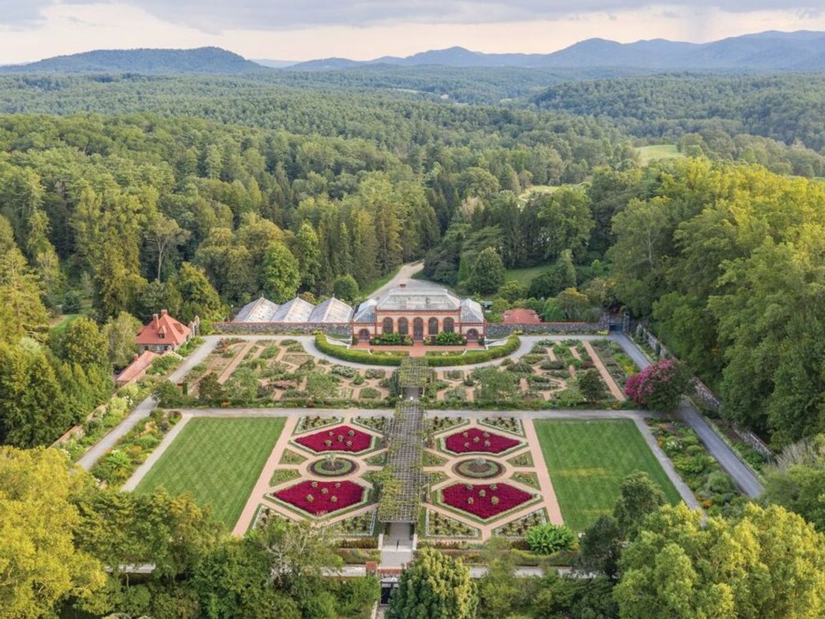 Aerial view of a grand historic estate surrounded by manicured gardens, lush trees, and mountain scenery under a partly cloudy sky