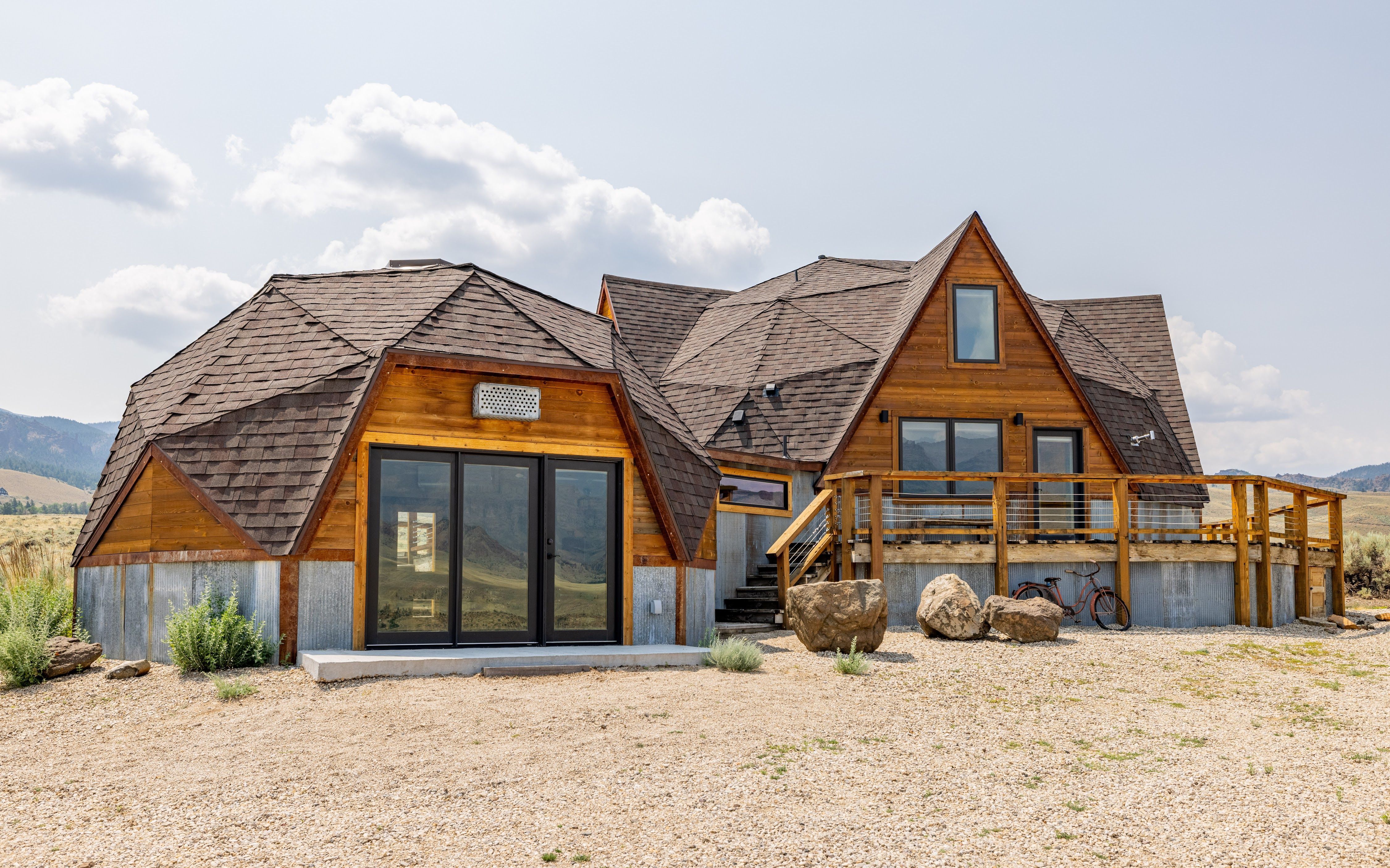 Dome Home in Cody and East Yellowstone features rounded shingle roofs, wood siding, and large glass doors. A raised deck overlooks open grassland with mountains in the distance.