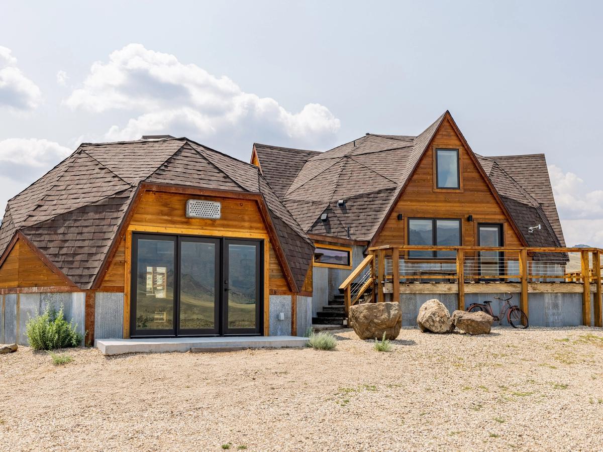 Dome Home in Cody and East Yellowstone features rounded shingle roofs, wood siding, and large glass doors. A raised deck overlooks open grassland with mountains in the distance.