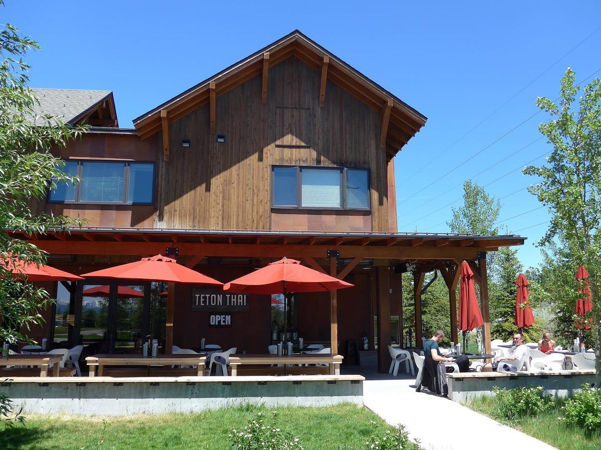 The timber-framed exterior of Teton Thai stands beneath a brilliant blue sky, its covered patio lined with bright red umbrellas and communal wooden picnic tables. Guests relax in the shade near the restaurant's "Open" sign on a sunny summer afternoon.