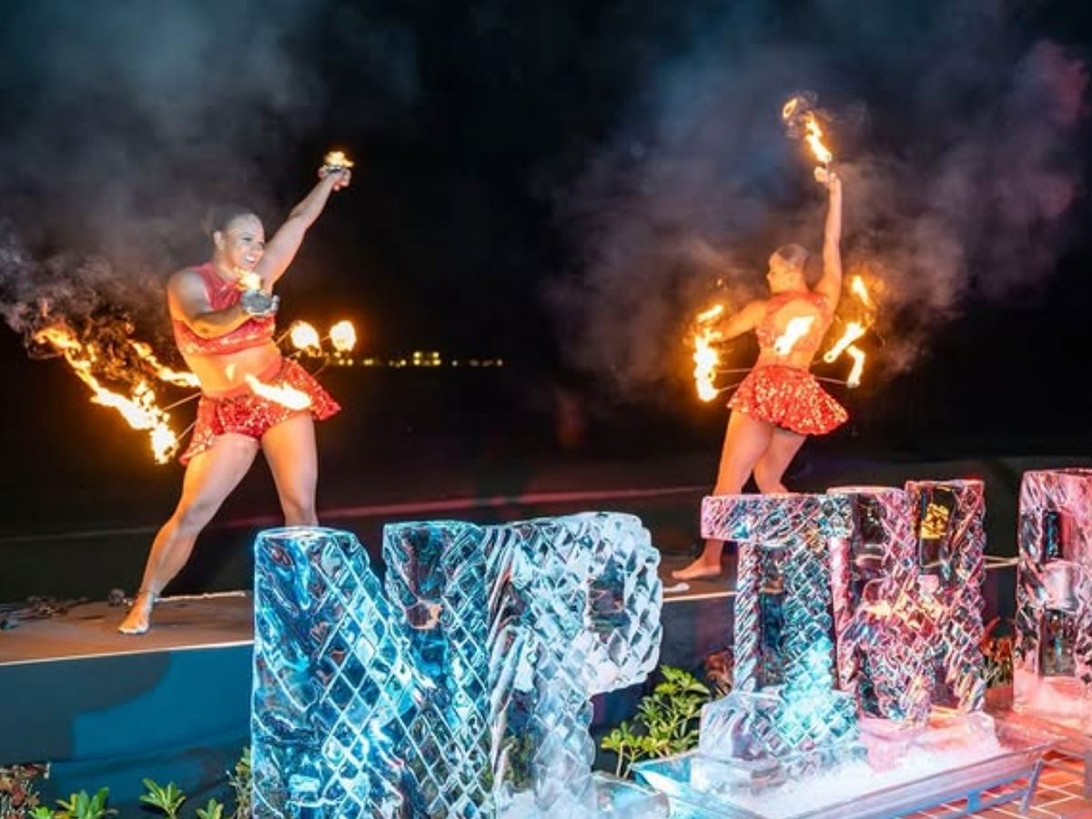 Two performers in red sequined outfits spin flaming batons at night in front of a glowing ice sculpture that reads “NTP,” with smoke and dramatic lighting adding to the high-energy show.
