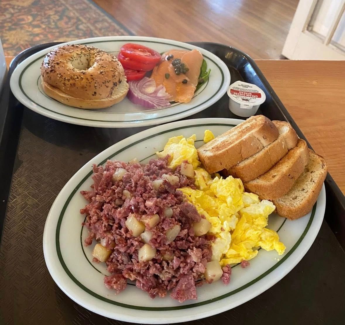 A classic breakfast plate with scrambled eggs, corned beef hash, toast, and a side of smoked salmon with bagel toppings. The hearty meal and casual setup capture the comfort of a small-town morning café.