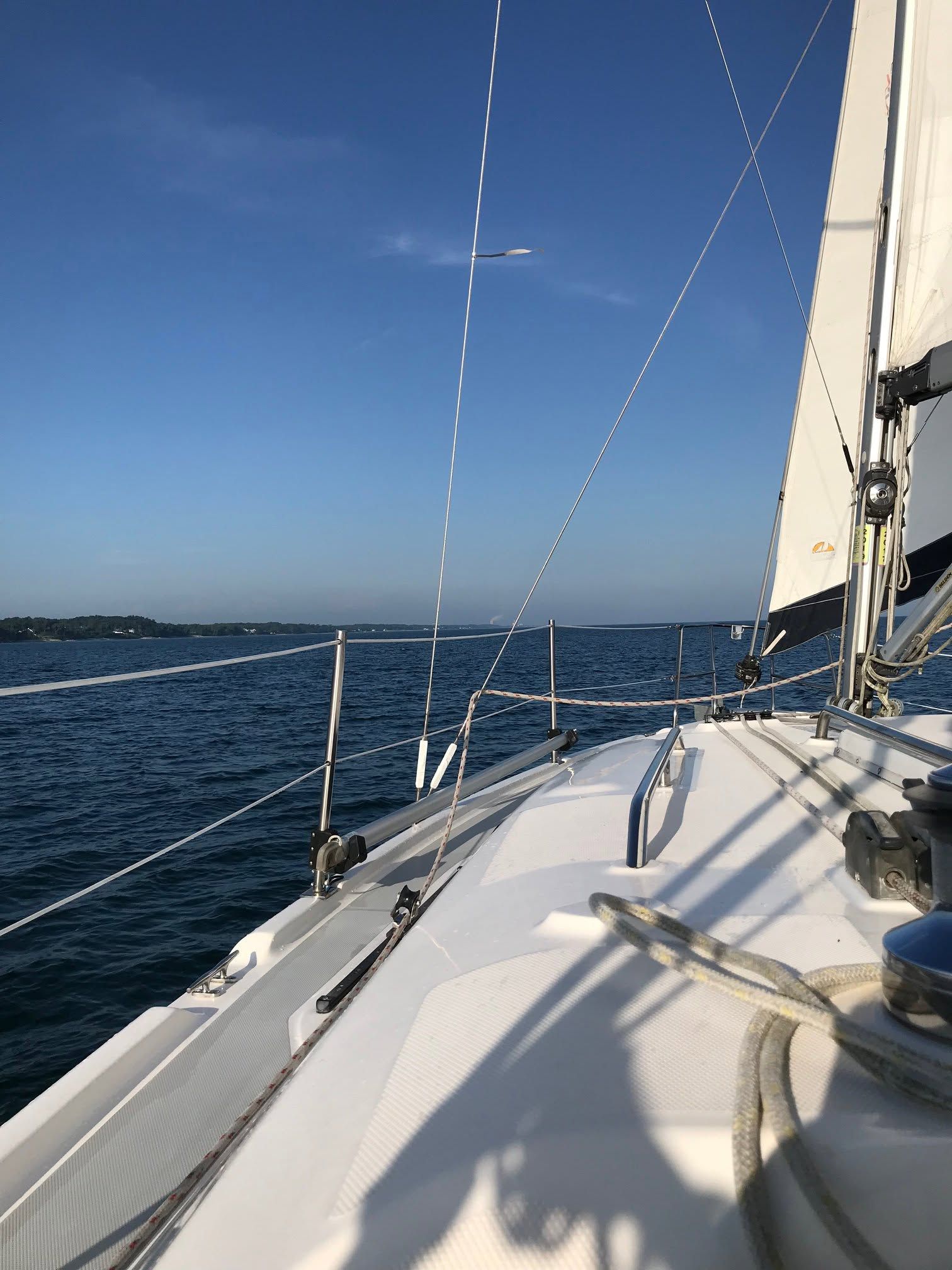  The view from the deck of a sailboat looks out over deep blue water toward a distant tree-lined shore, with rigging and a white sail rising against a clear sky. Sunlight casts crisp shadows across the boat's bow on a perfect day for sailing.