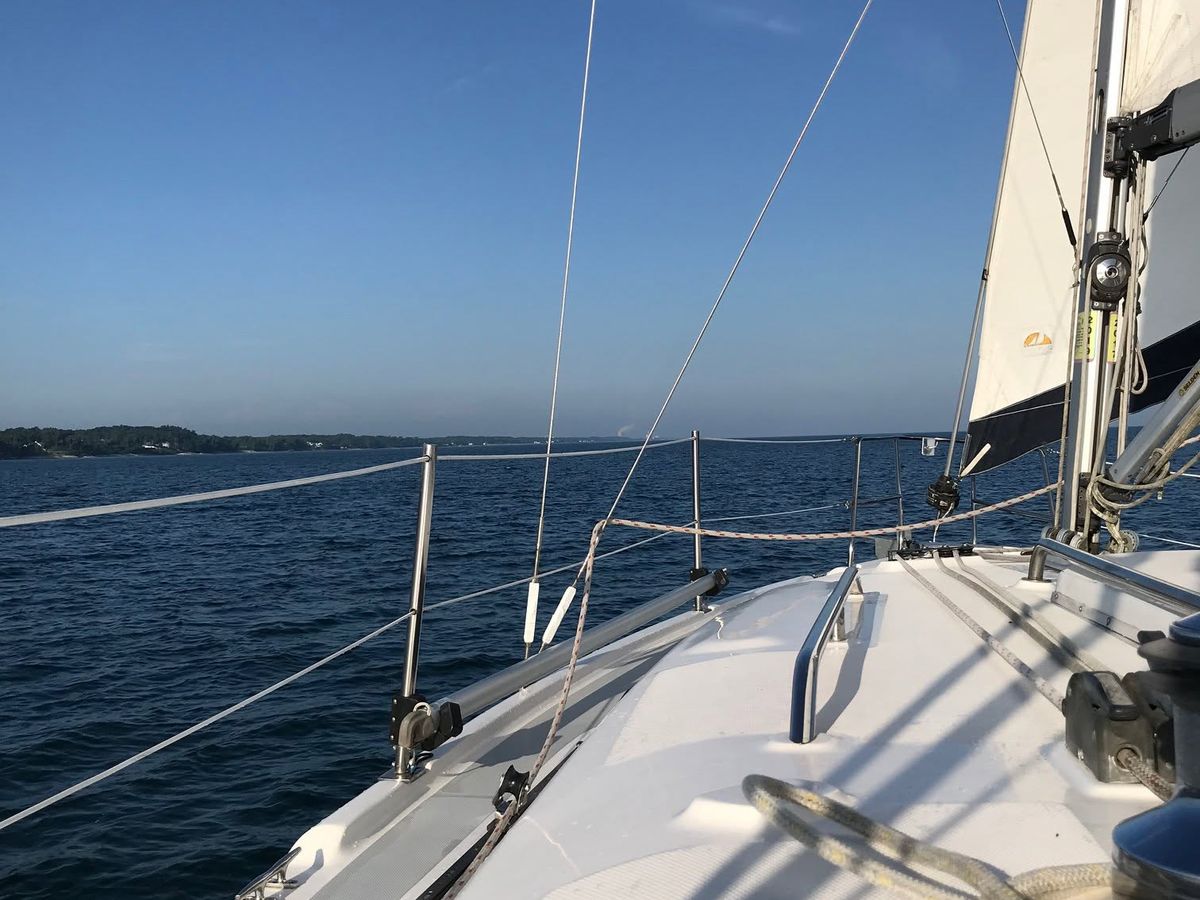 The view from the deck of a sailboat looks out over deep blue water toward a distant tree-lined shore, with rigging and a white sail rising against a clear sky. Sunlight casts crisp shadows across the boat's bow on a perfect day for sailing.