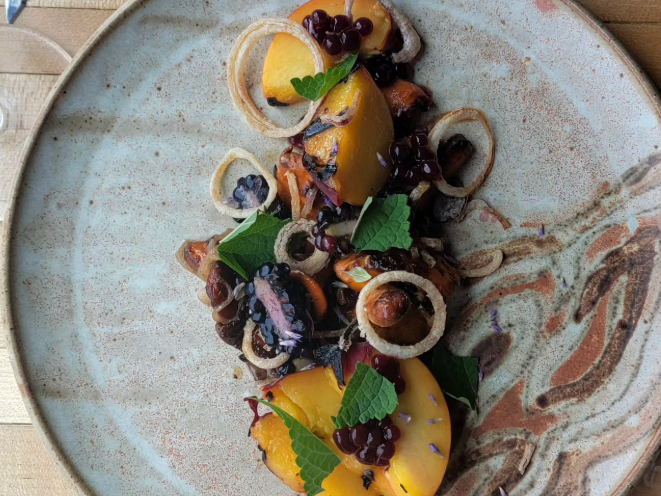 A plated dish featuring sliced peaches, blackberries, fried onion rings, herbs, and garnishes arranged artfully on a ceramic plate.