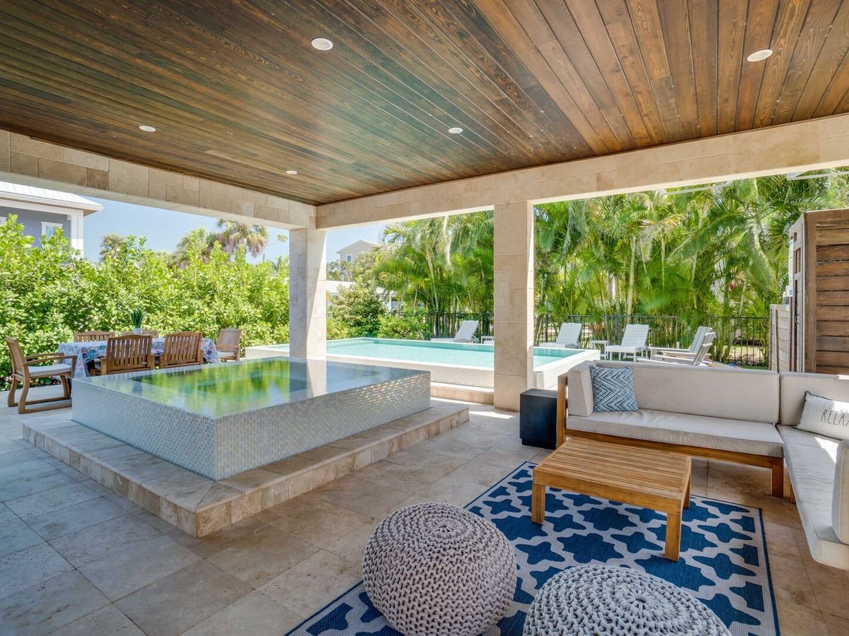 Covered outdoor living space with a wood-paneled ceiling, lounge seating, and a raised reflecting pool overlooking a private swimming pool framed by lush tropical greenery.