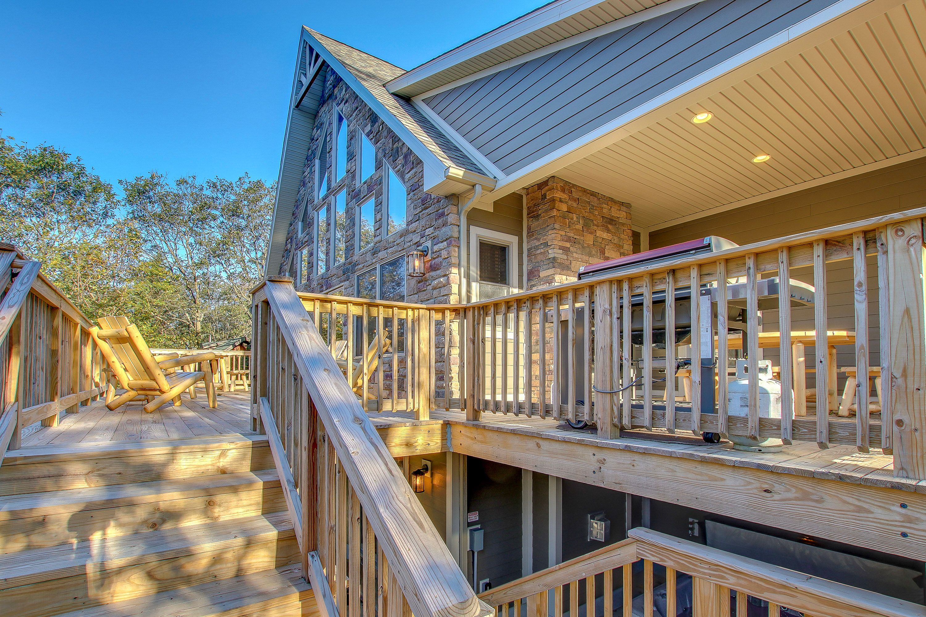 Wooden staircase and deck leading to a rustic cabin with large triangular windows, stone exterior, and a grill area under a covered section.