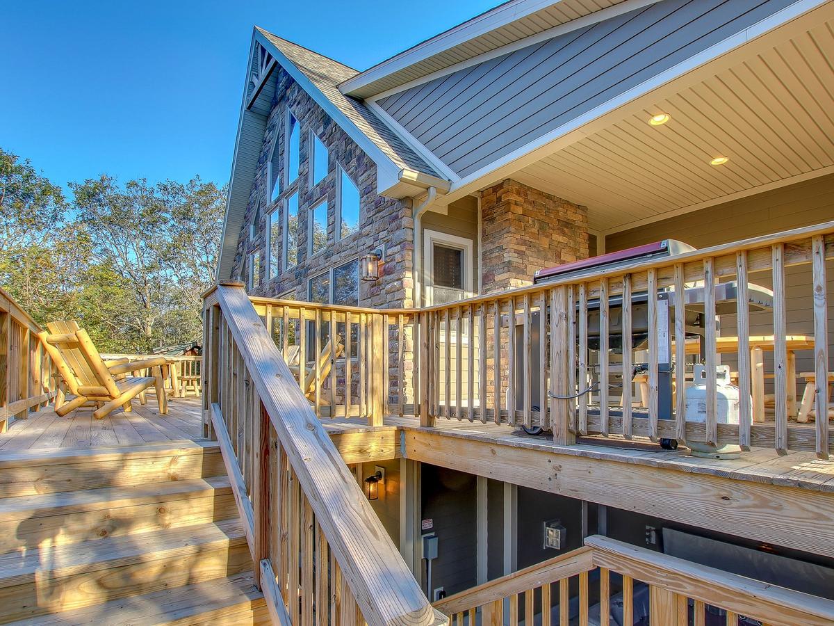 Wooden staircase and deck leading to a rustic cabin with large triangular windows, stone exterior, and a grill area under a covered section.