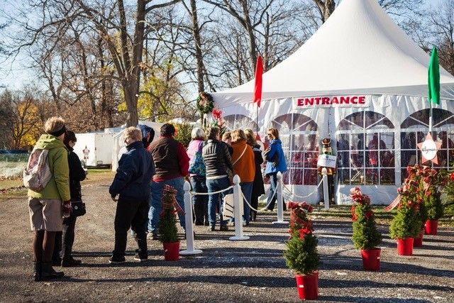 Visitors bundled in winter coats line up at the entrance tent of the Canandaigua Christkindl Market in upstate New York, with potted Christmas trees and festive red planters lining the pathway on a crisp autumn day