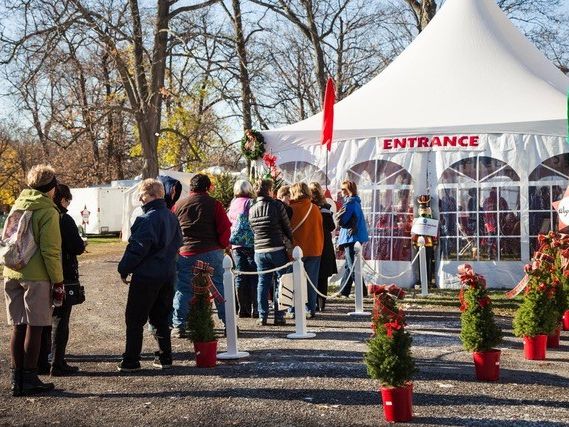 Visitors bundled in winter coats line up at the entrance tent of the Canandaigua Christkindl Market in upstate New York, with potted Christmas trees and festive red planters lining the pathway on a crisp autumn day