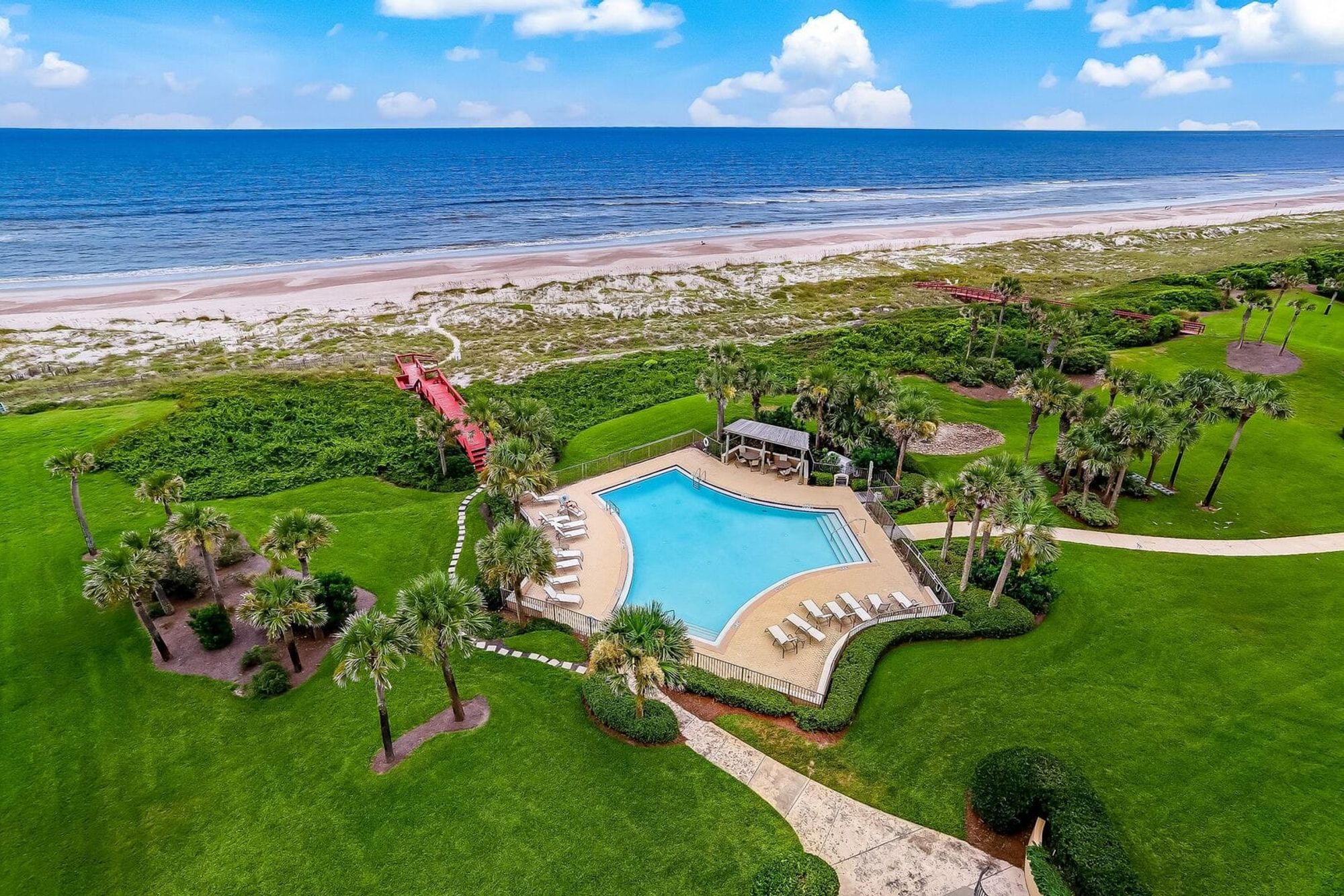 Aerial shot of a beachfront resort with a large pool, palm trees, and a red boardwalk leading to the shore. Blue skies and clear ocean make it the ideal beach vacation spot.