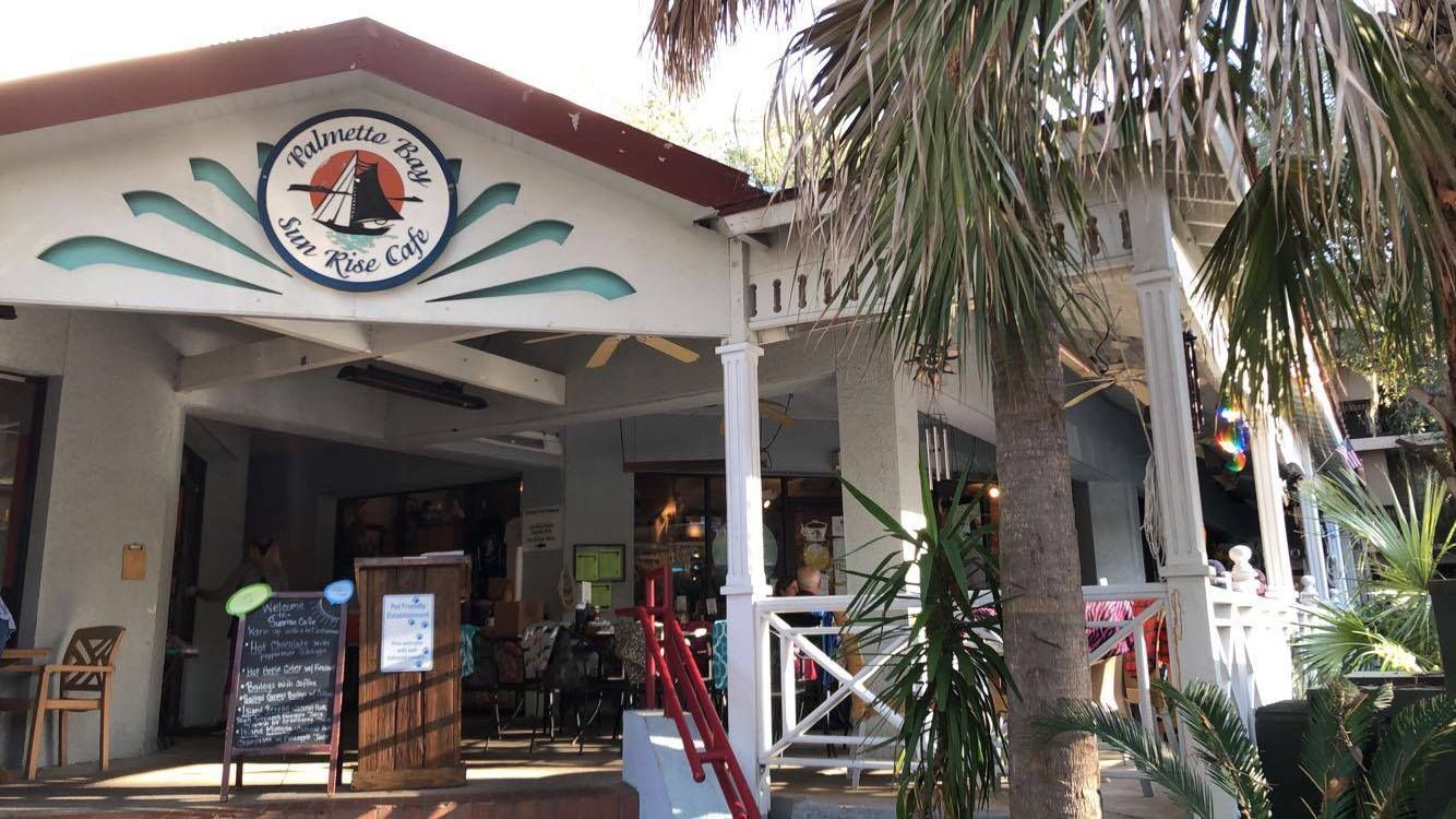 The entrance of Palmetto Bay Sunrise Cafe, a cozy local breakfast spot surrounded by palm trees and coastal charm. Guests gather on the shaded patio to enjoy coffee and classic Southern breakfast dishes. This popular Hilton Head cafe is known for relaxed island vibes and fresh morning favorites.